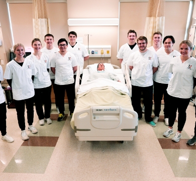 nursing students stand around a hospital bed with a manikin in it.